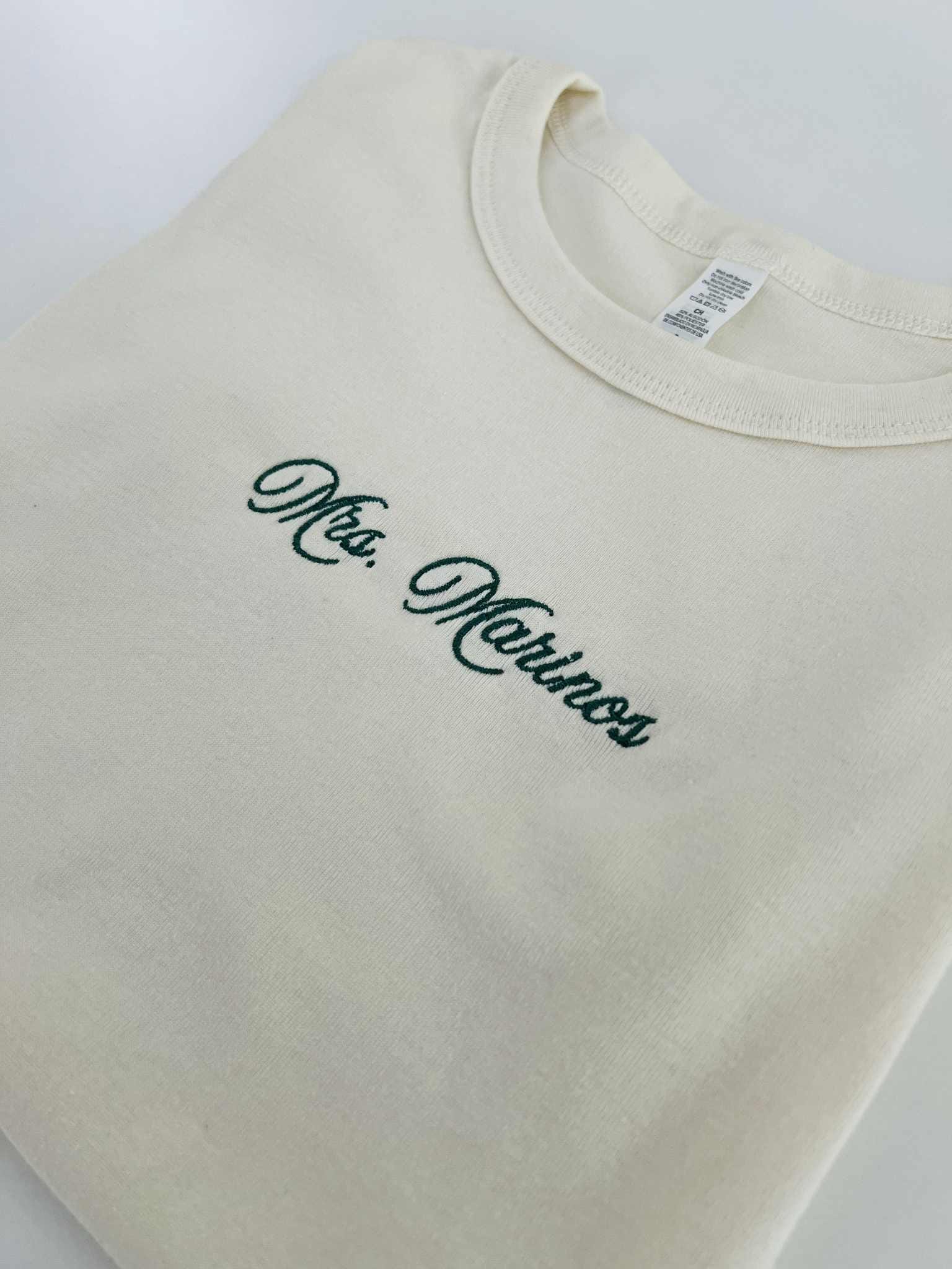 Close-up of Mrs. Marinos embroidery on a cream baby tee