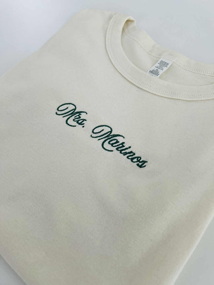 Close-up of Mrs. Marinos embroidery on a cream baby tee